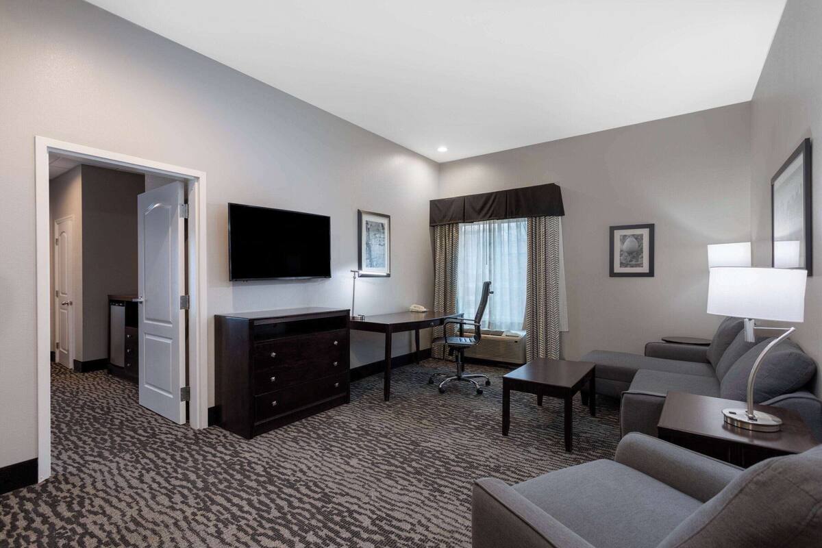 A hotel room with a flat-screen TV, a desk with a chair, a sofa, a lamp, and a large window with curtains.