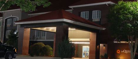 A hotel entrance with a covered driveway, a parking area, and a sign with the hotel's name.
