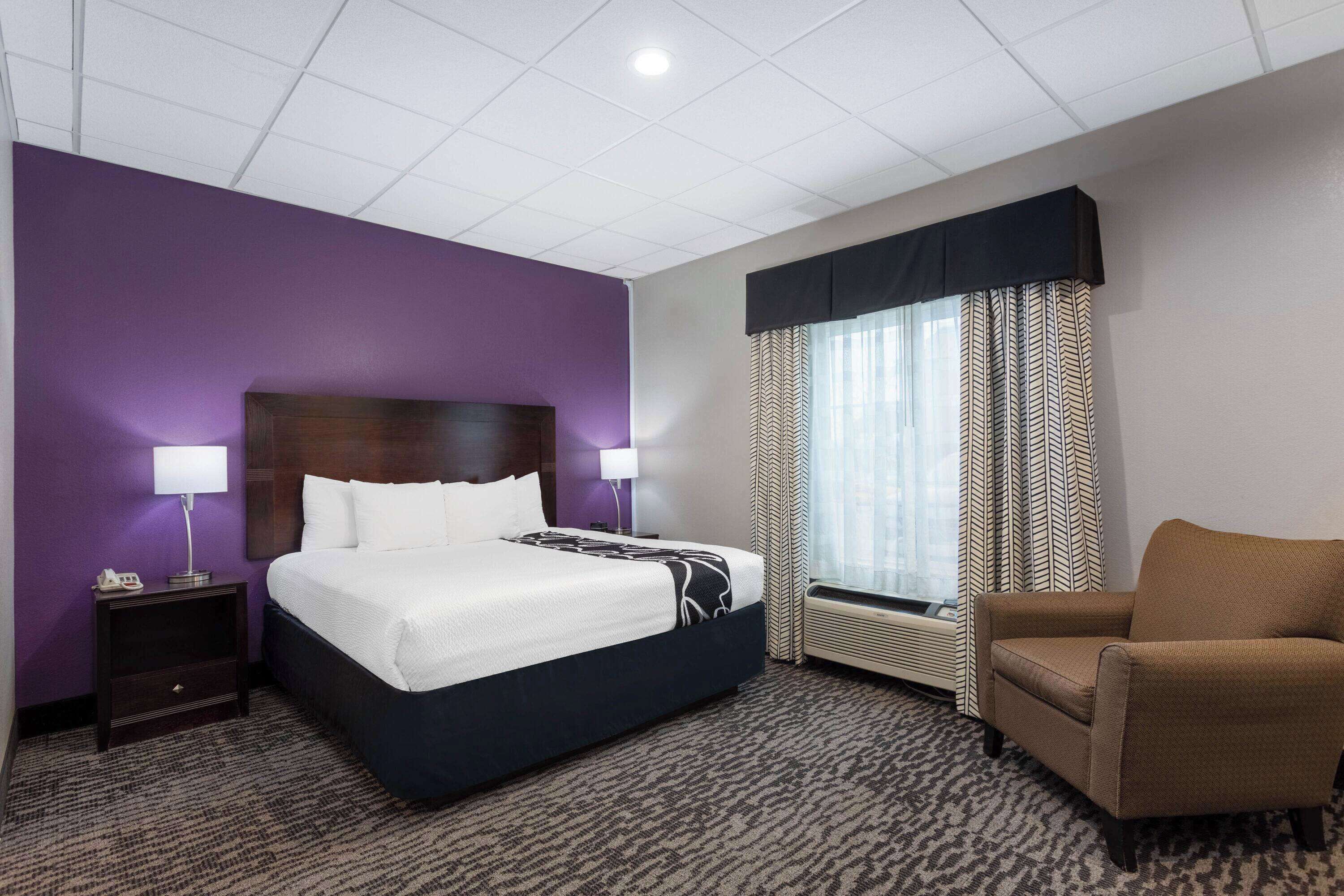 A hotel room with a bed, a chair, a nightstand, a lamp, a window with curtains, and a wall with a purple accent.