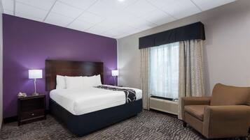 A hotel room with a bed, a chair, a nightstand, a lamp, a window with curtains, and a wall with a purple accent.