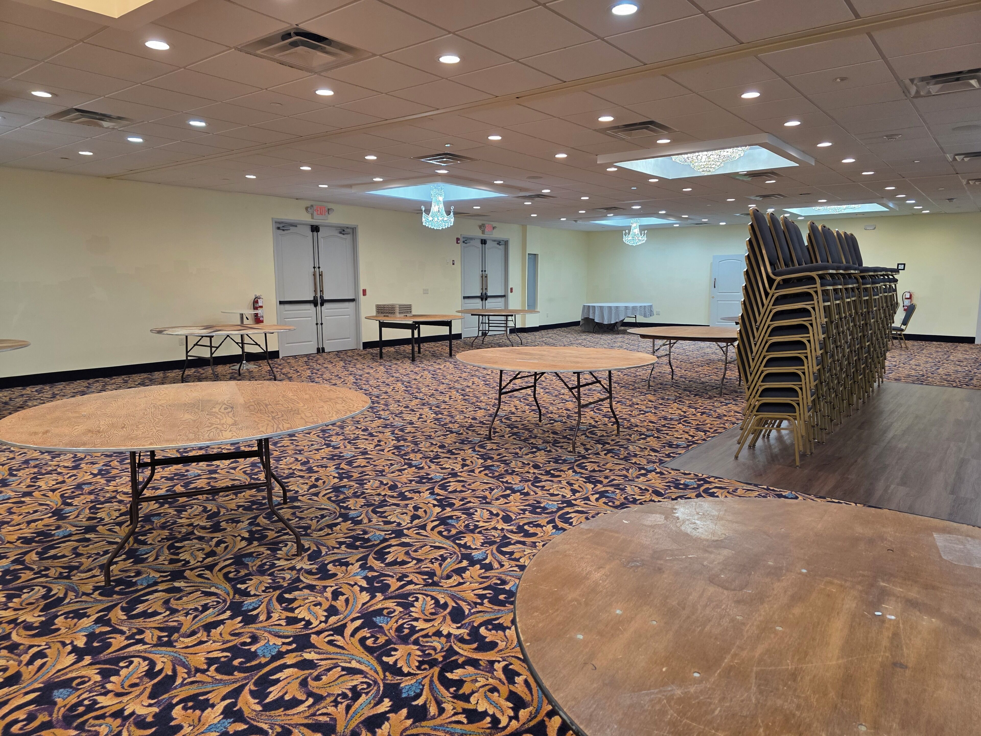 Habitación de hotel con alfombra estampada, mesas redondas y una pila de sillas.