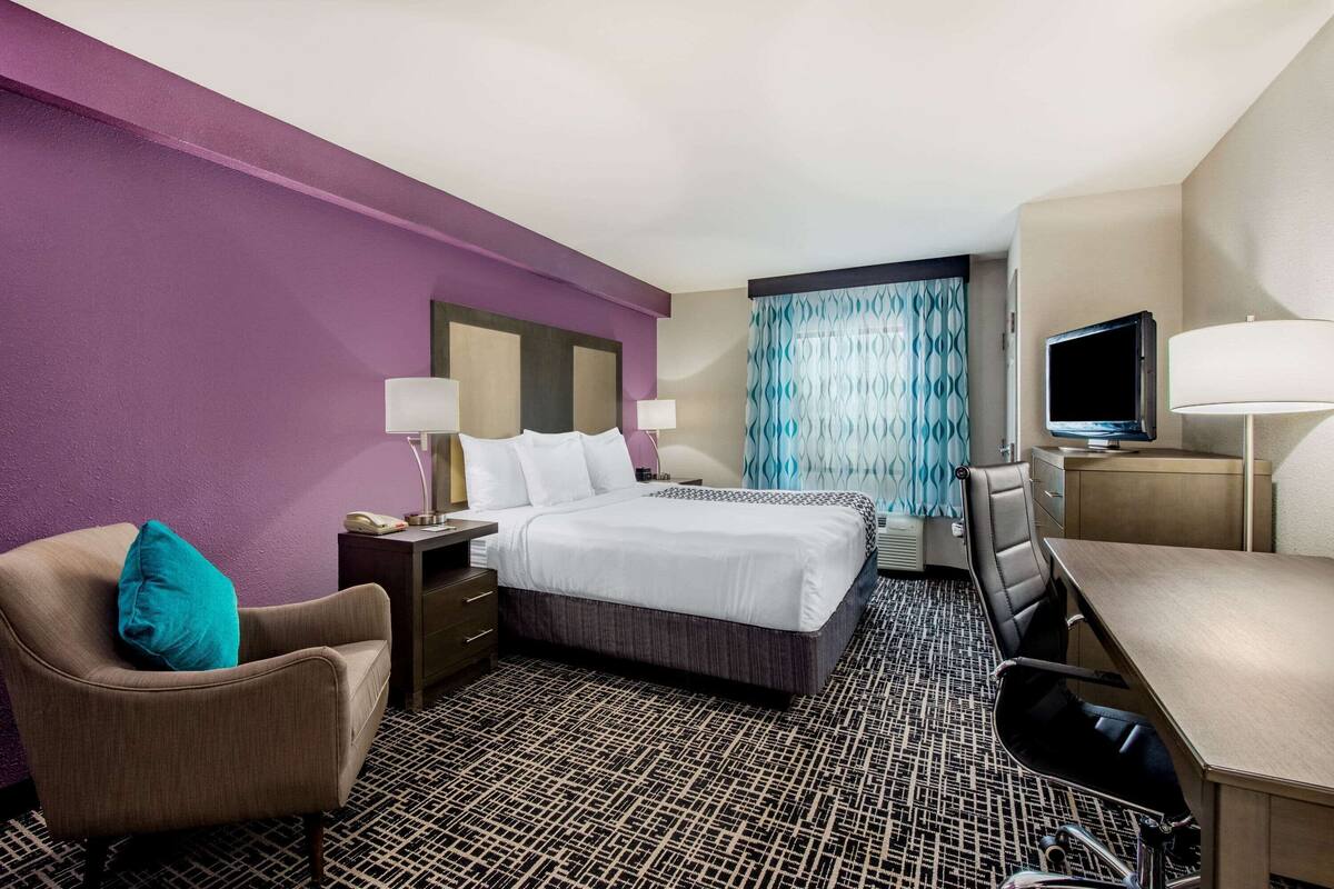 Une chambre d’hôtel avec un lit, un bureau, une chaise, une télévision et une fenêtre avec des rideaux à motifs.