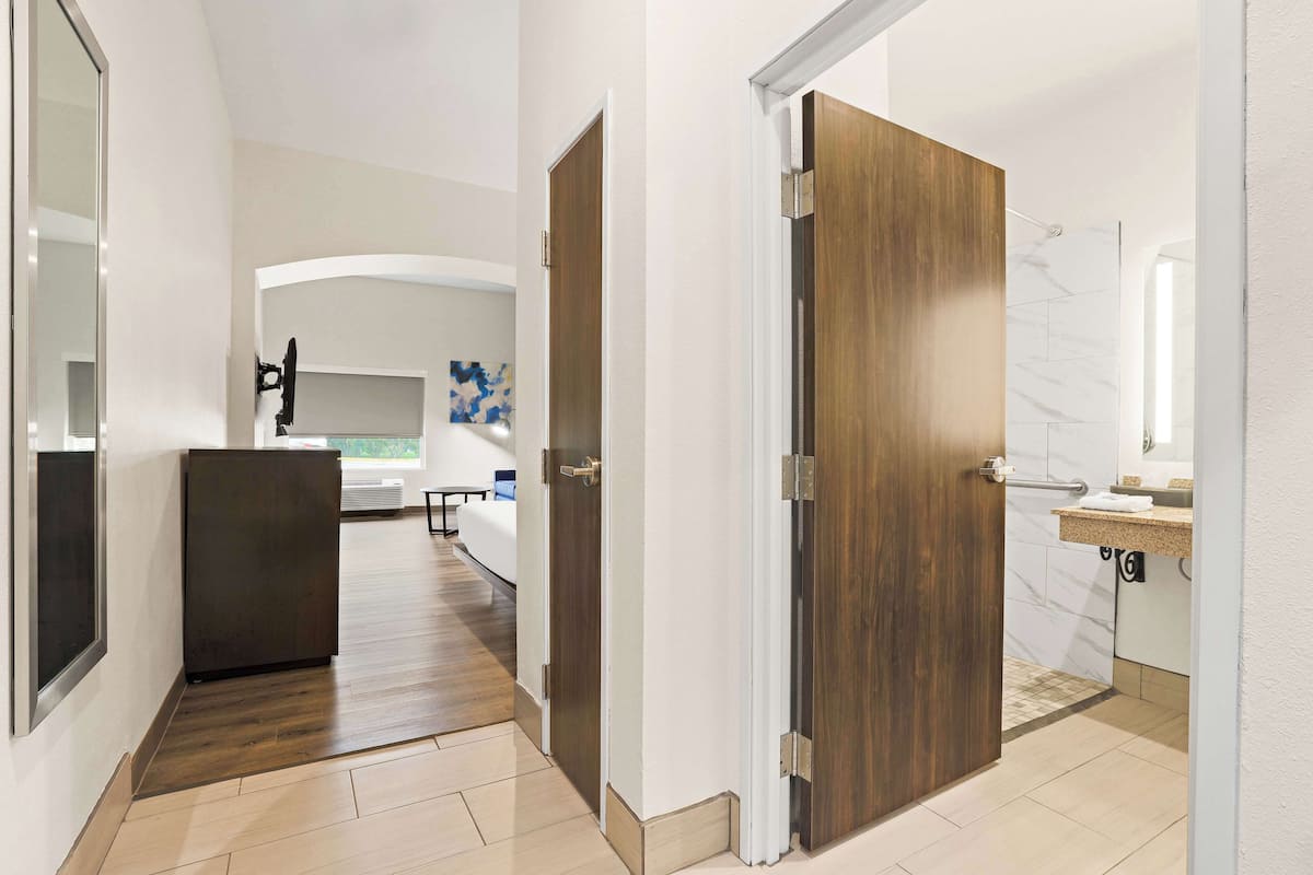 A hotel hallway with a wooden door, a small refrigerator, and a bathroom with a marble wall and sink.