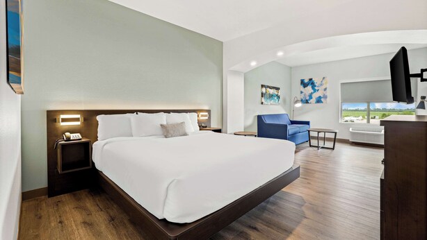Habitación de hotel con una cama grande, una mesita de noche con un teléfono, una pequeña mesa, una silla azul y un ventanal con vistas.