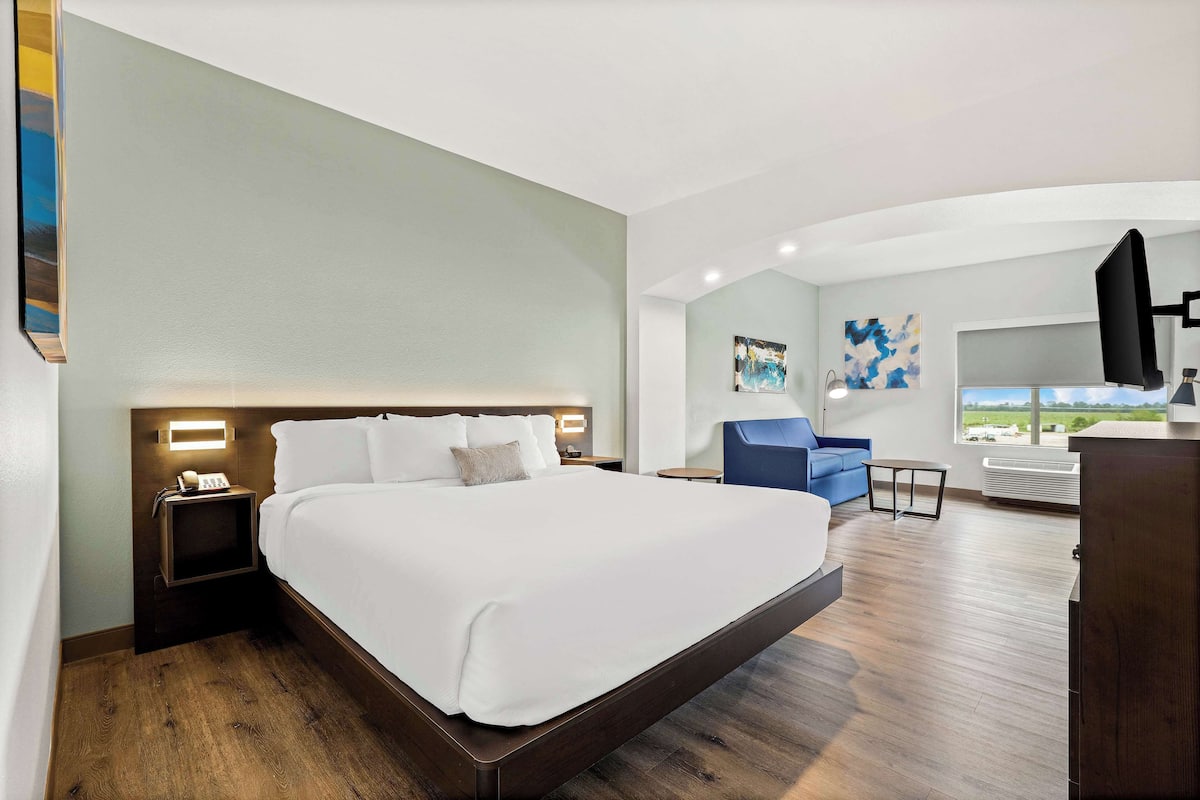 A hotel room with a large bed, a bedside table with a phone, a small table, a blue chair, and a large window with a view.