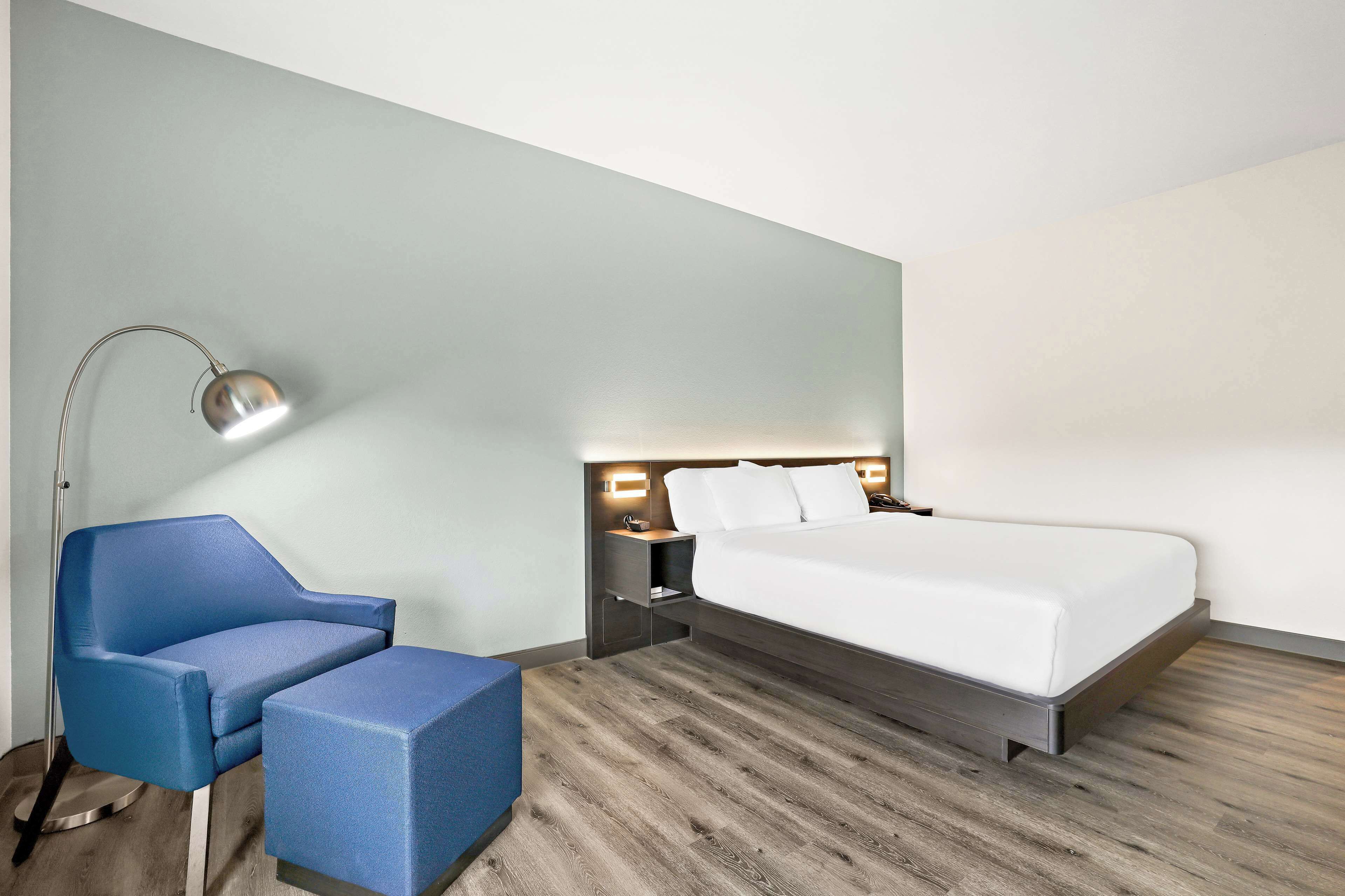 A modern hotel room with a bed, a blue armchair, a side table, and a floor lamp.