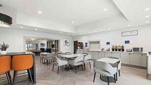 A modern dining area with multiple tables and chairs, a kitchen area with appliances, and a sign that reads "Come gather at our table."
