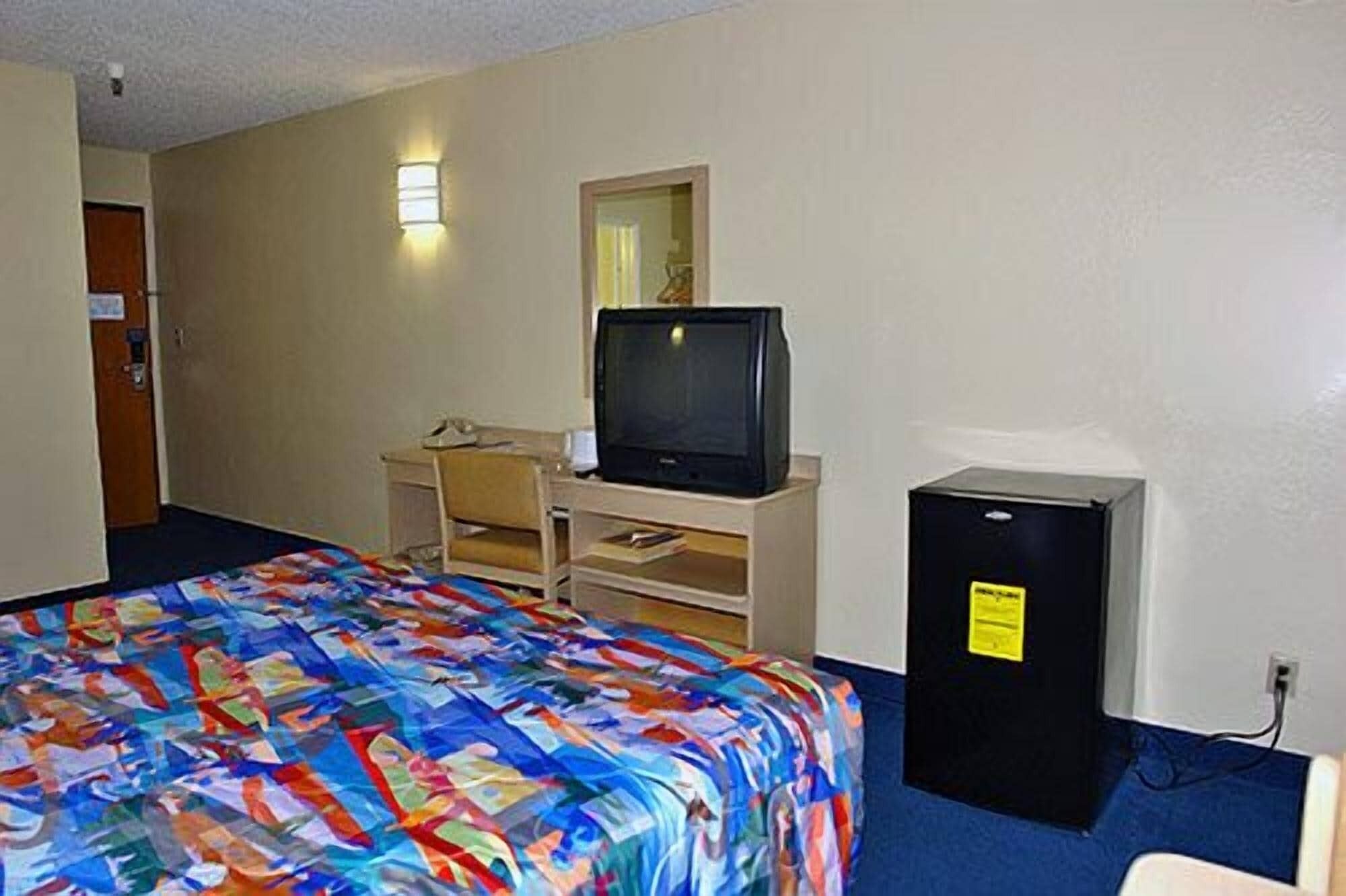 Una camera d'albergo con un letto, una scrivania con un computer, una televisione e un piccolo frigorifero.
