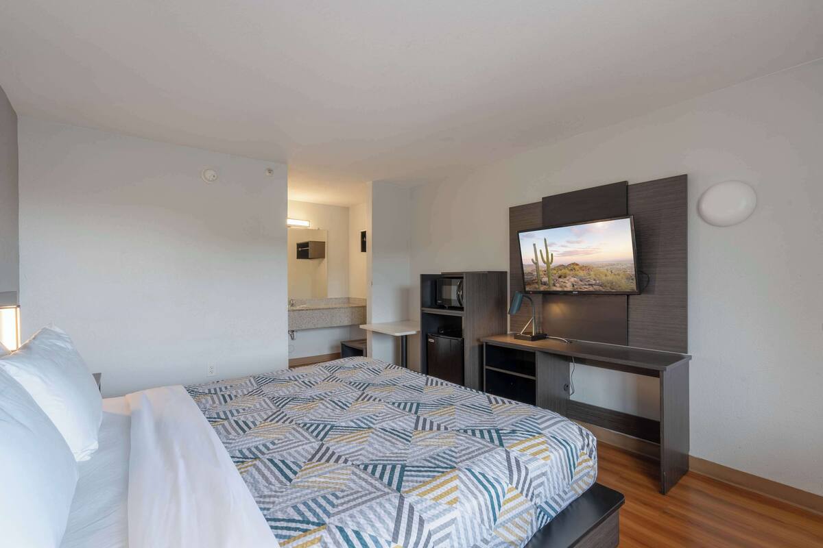 Una habitación de hotel moderna con una cama, un escritorio con televisor de pantalla plana, un espejo y una pequeña kitchenette.