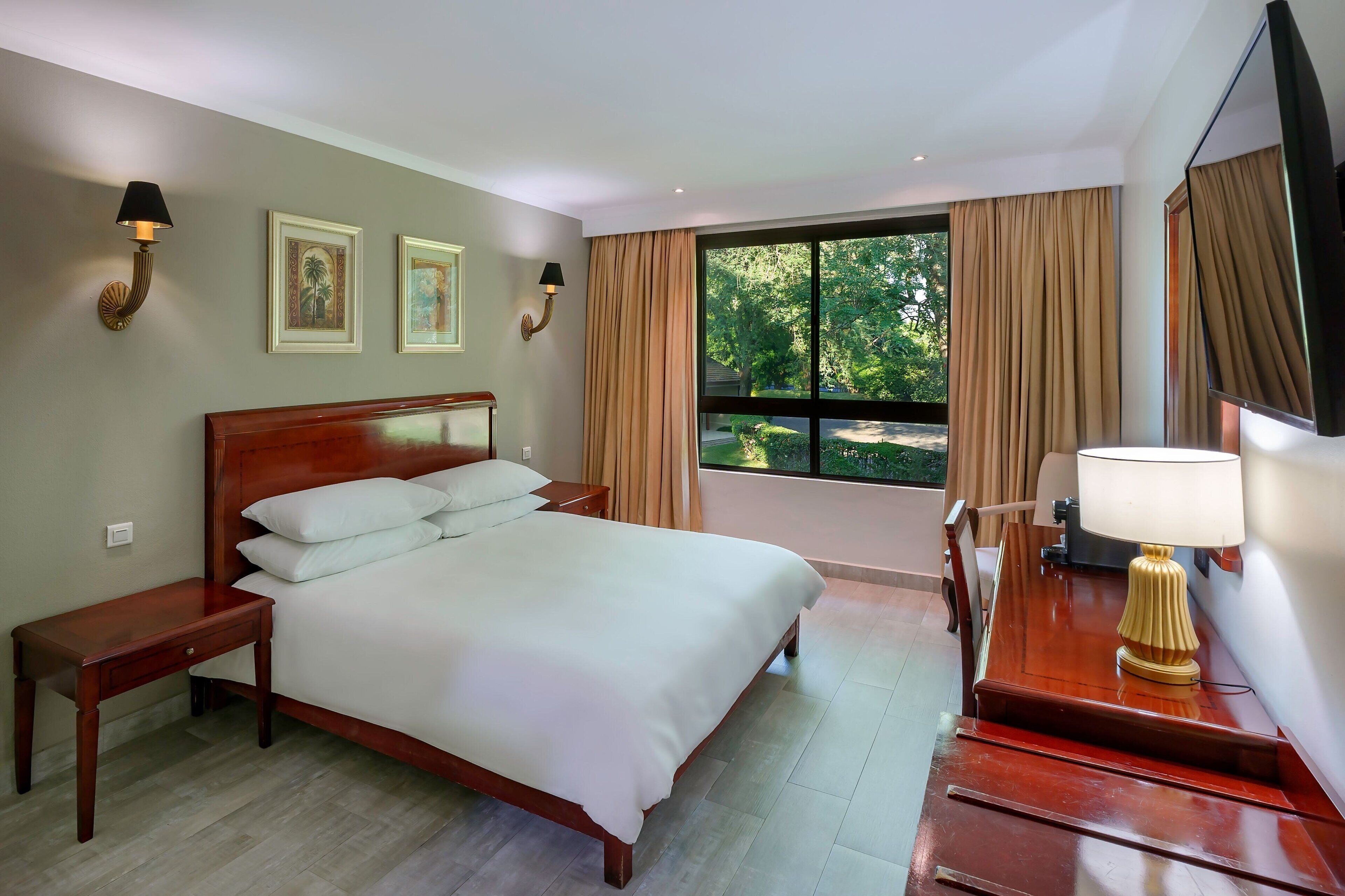 A neatly made bed with white linens, a wooden nightstand, a bedside table, a desk with a lamp, and a large window with curtains.