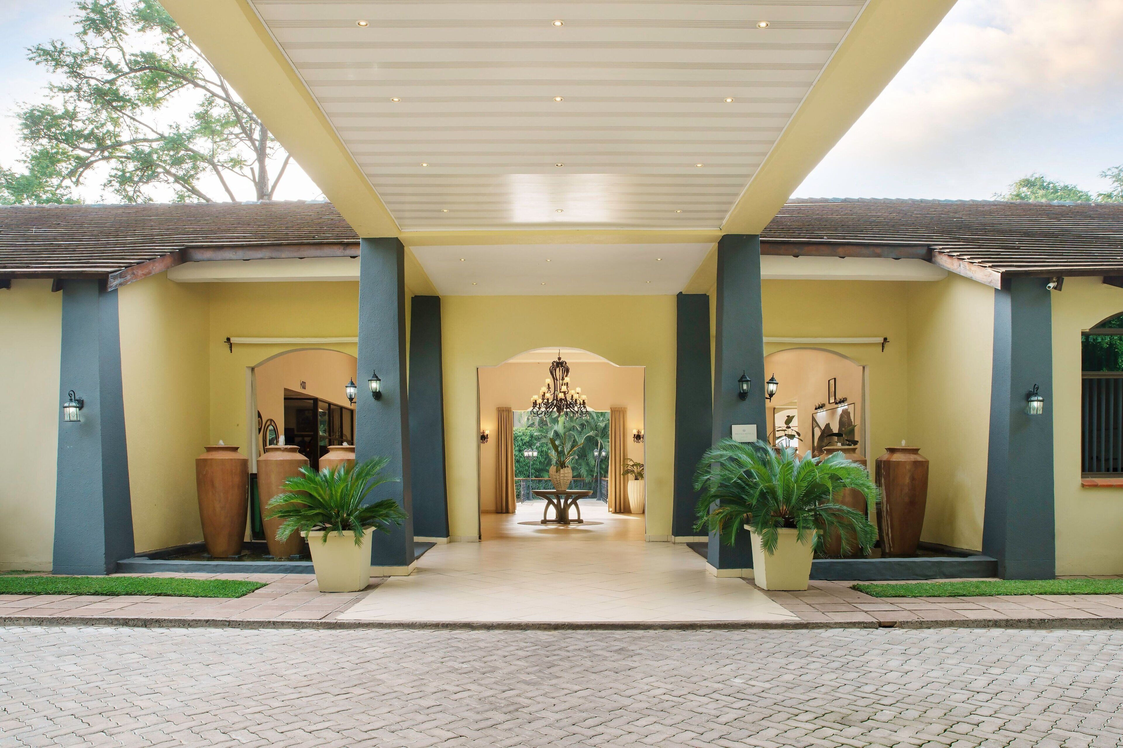 A hotel entrance with a covered walkway, large potted plants, and a chandelier.