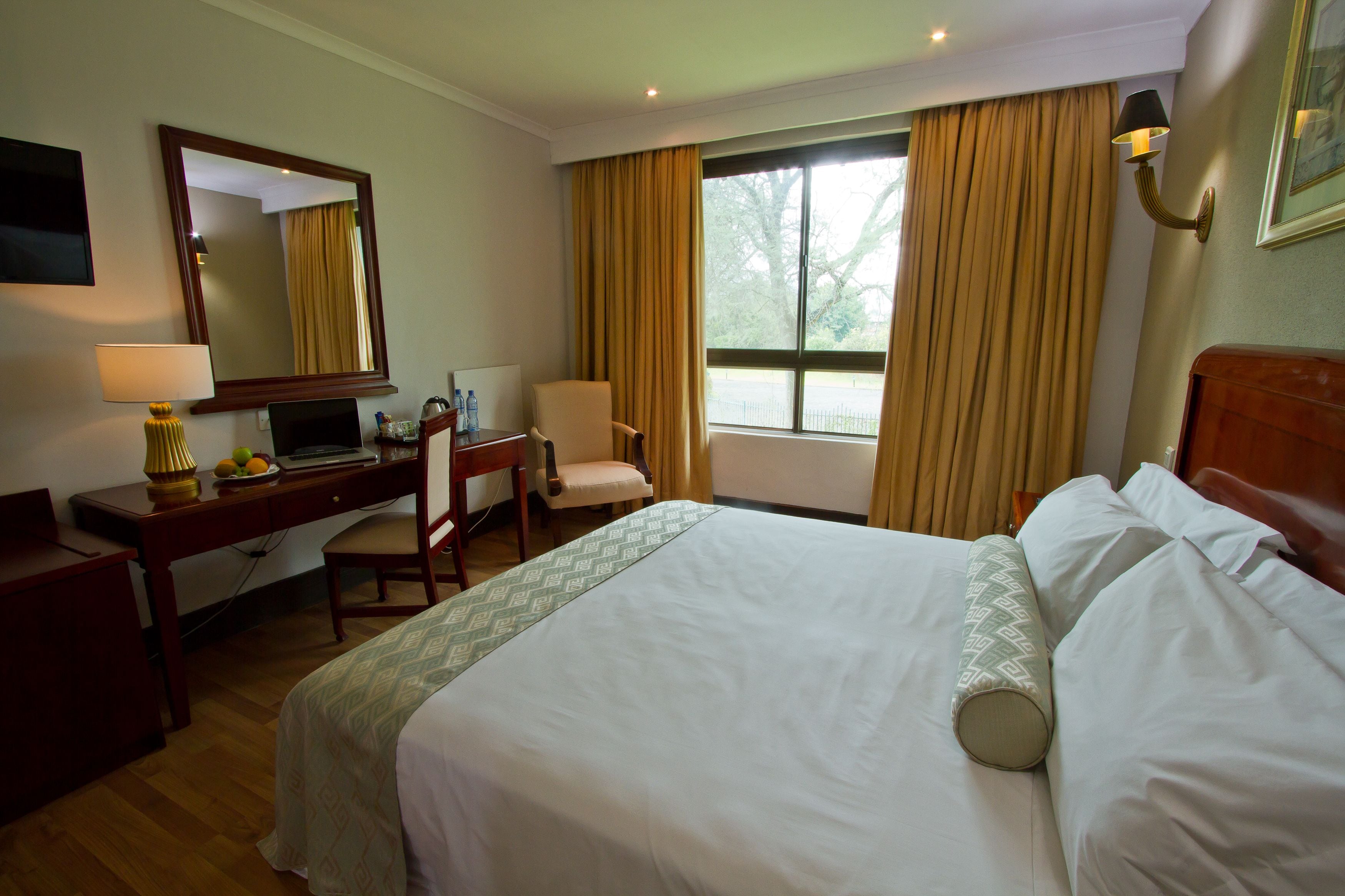 A hotel room with a bed, a desk with a lamp, a chair, and a large window with curtains.