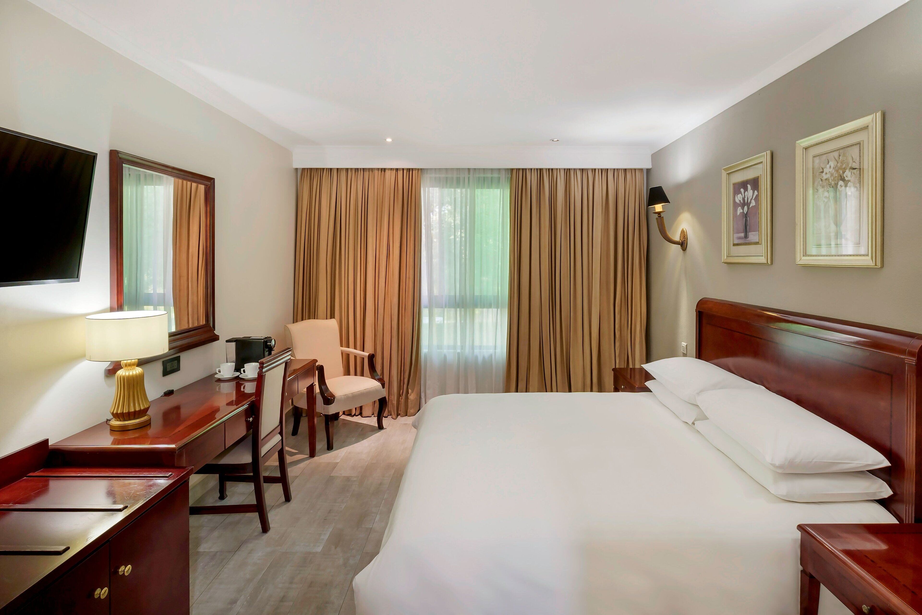 Ein Hotelzimmer mit einem Bett, einem Schreibtisch mit Lampe, einem Stuhl, einem Fernseher und Vorhängen.