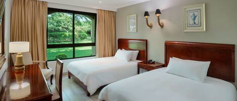 A hotel room with two beds, a desk with a lamp, a nightstand, and a view of a garden through a large window.