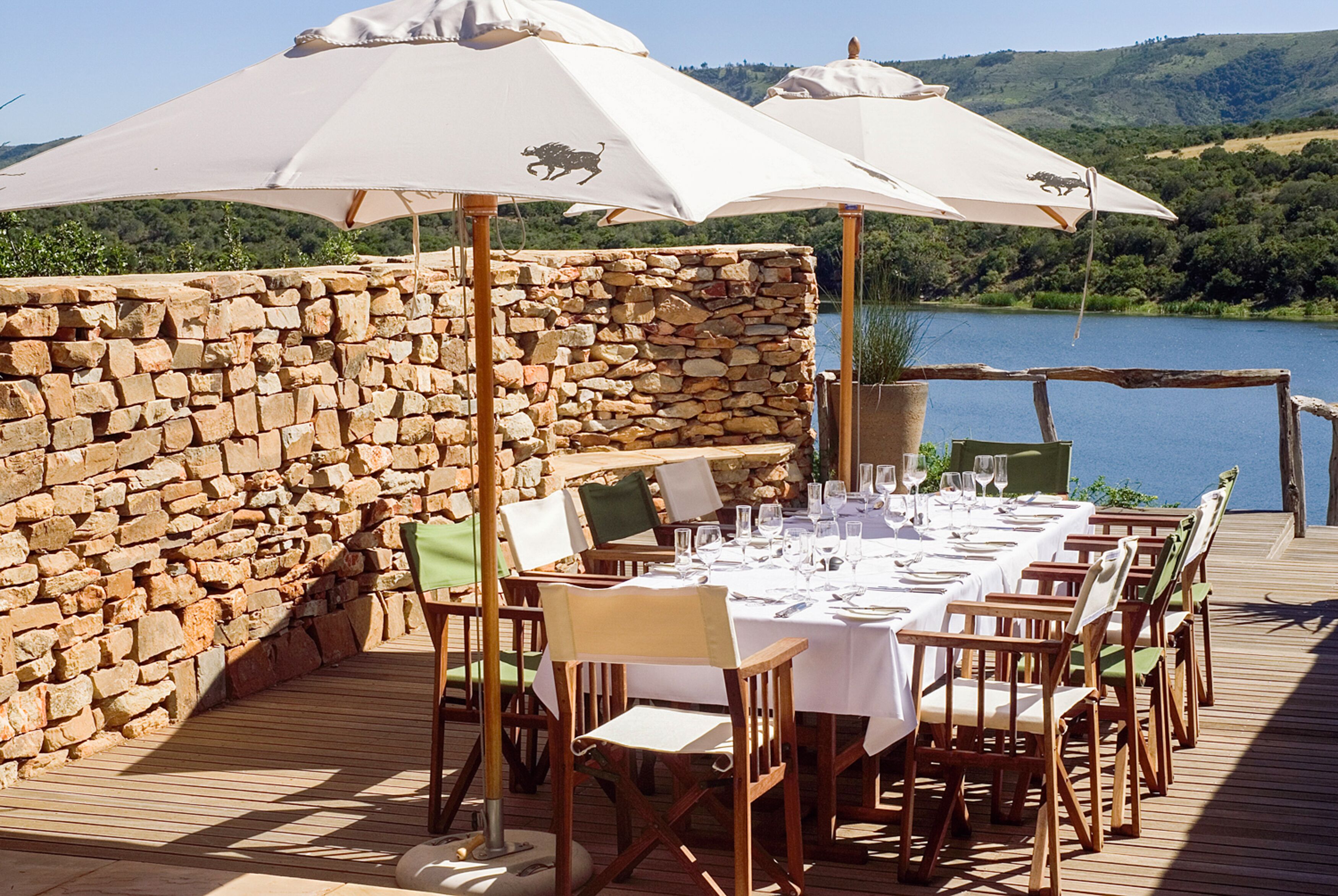 A table set for dining with white tablecloths and chairs, large umbrellas, and a stone wall backdrop.
