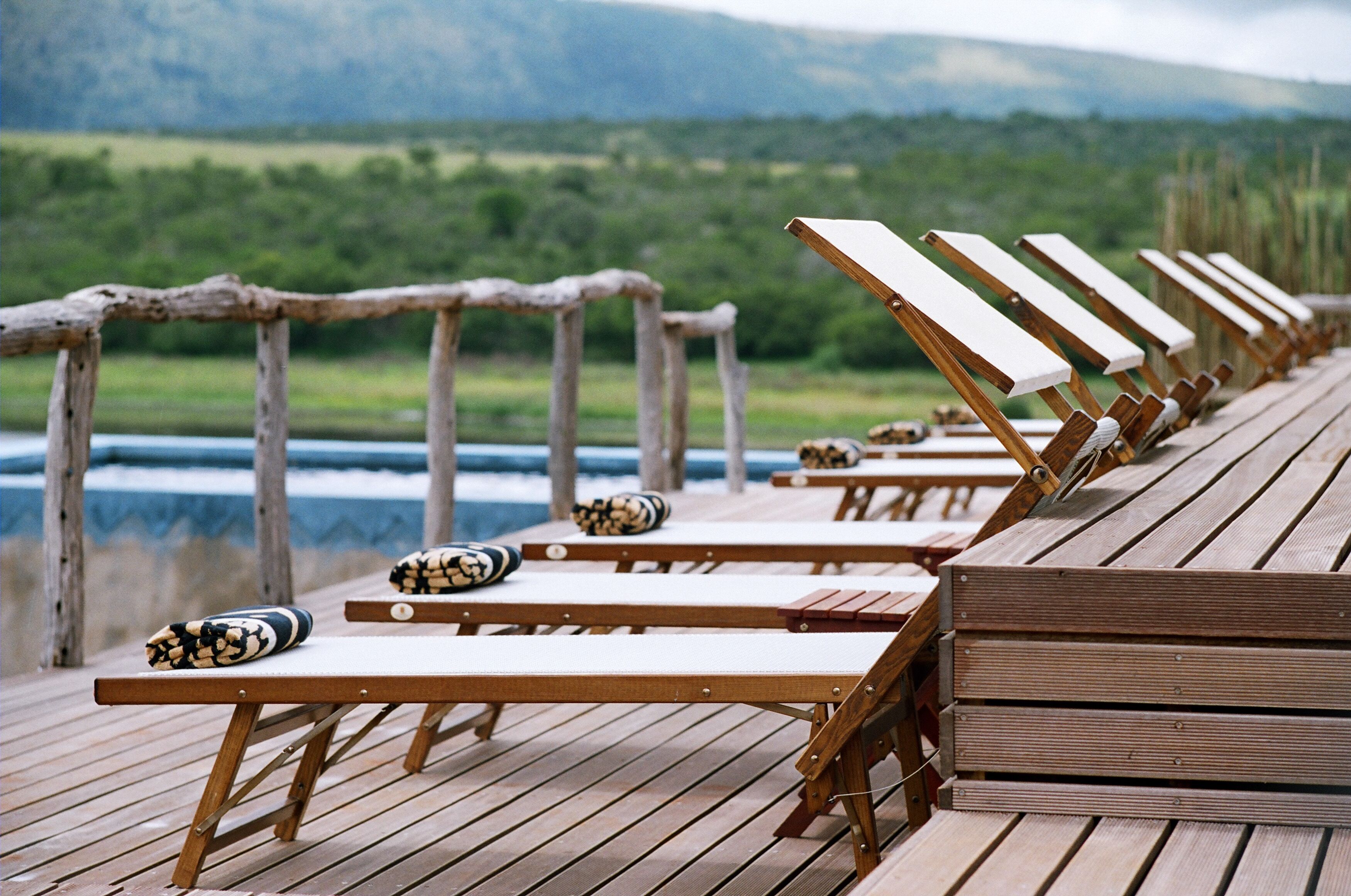 A wooden deck with lounge chairs and cushions, overlooking a body of water and greenery.