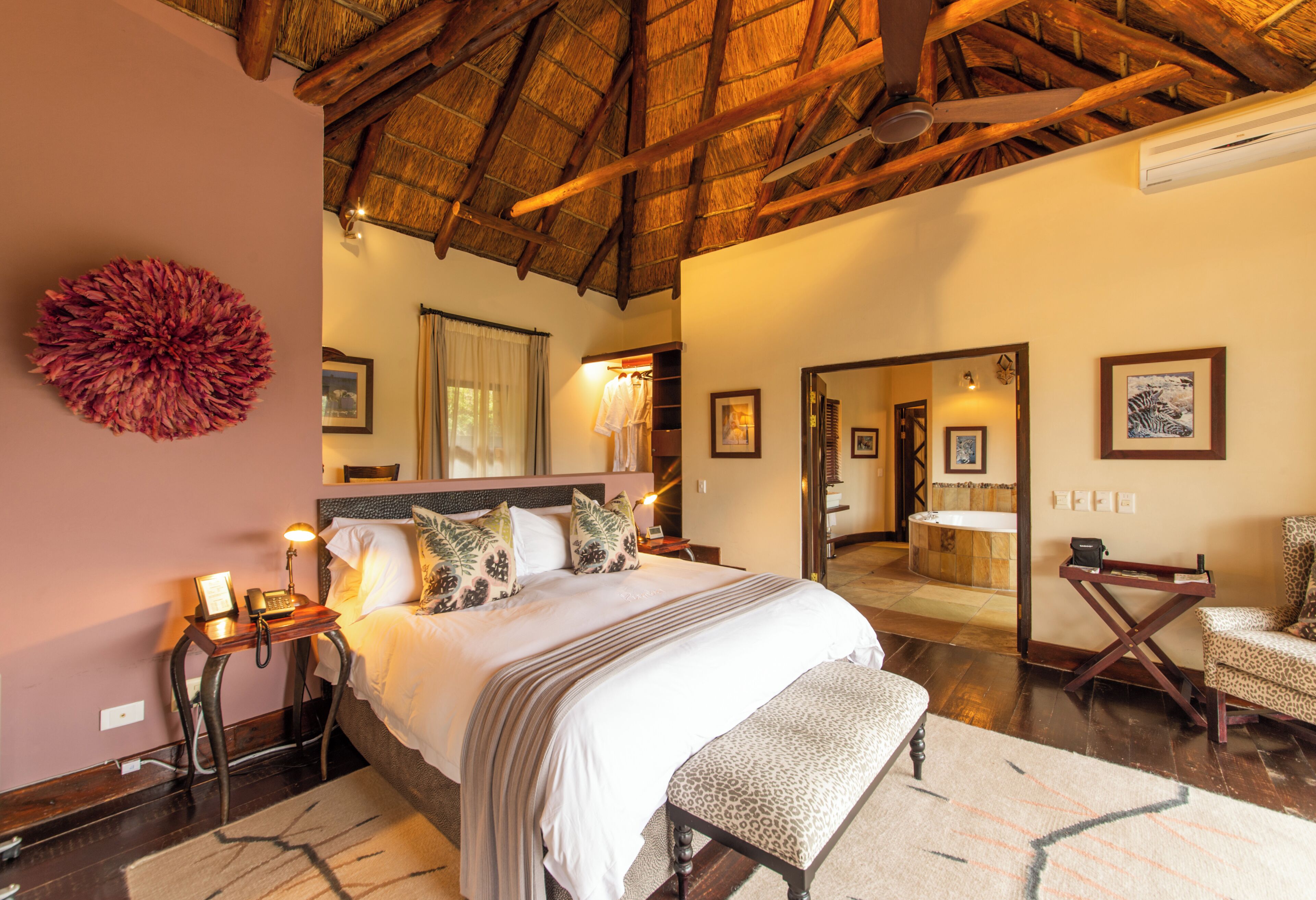 A spacious bedroom with a large bed, a wooden ceiling, a sitting area with a chair, and a bathroom visible through a doorway.