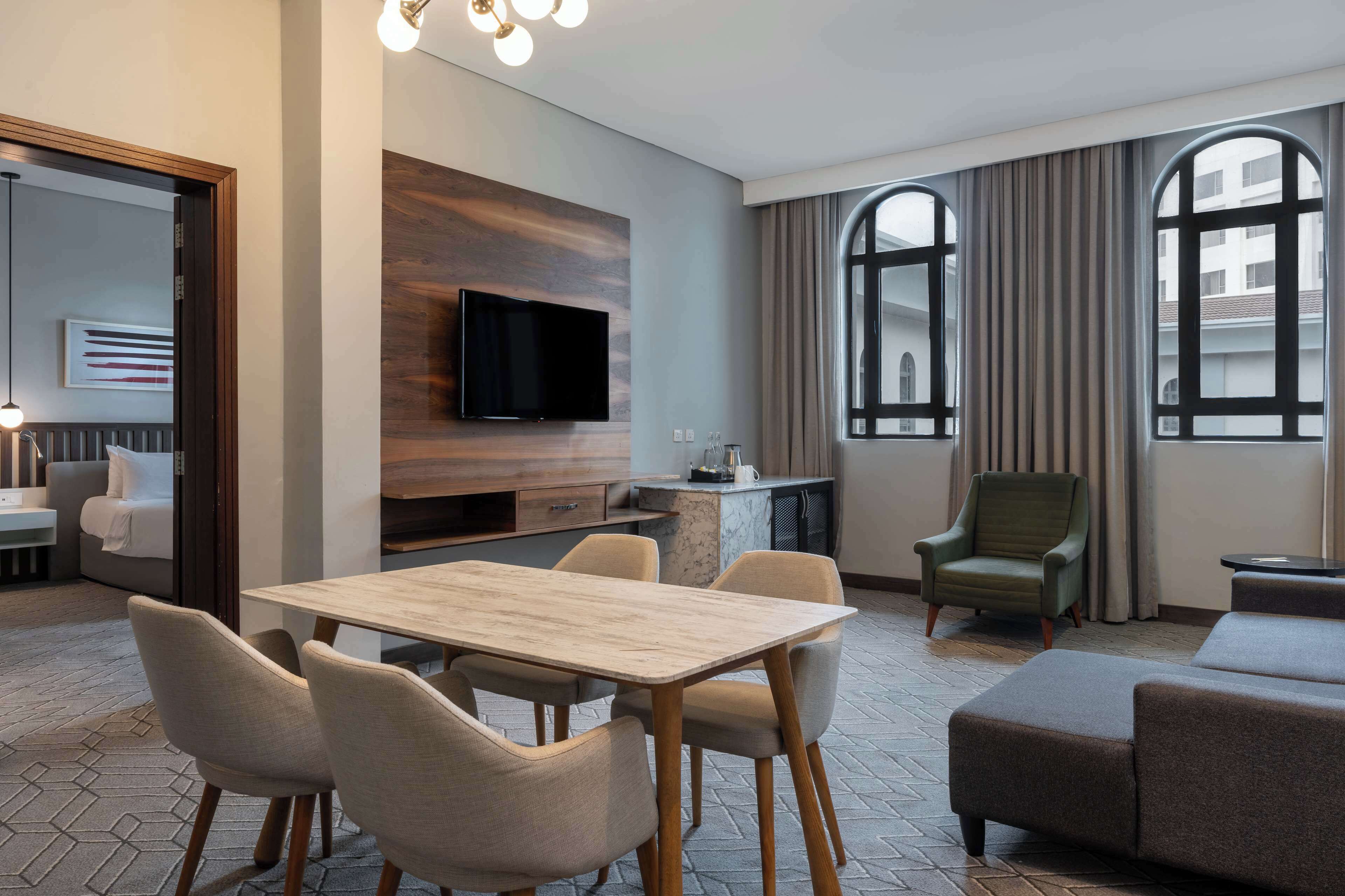 A modern hotel room with a dining area, a TV mounted on a wooden wall, a sofa, and large windows with curtains.