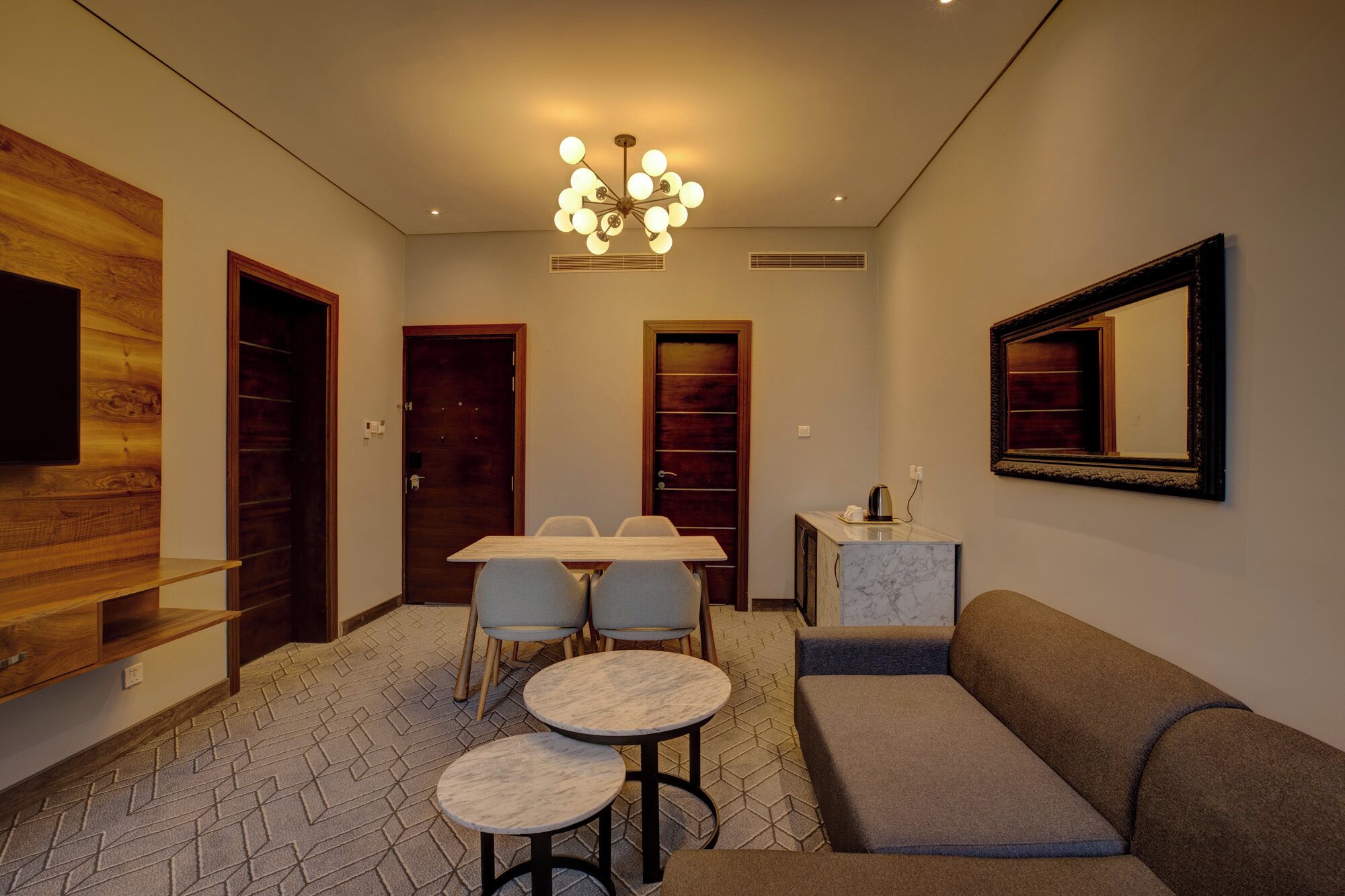 Een moderne woonkamer met een bank, twee ronde tafels, een eettafel met stoelen, een aan een houten paneel bevestigde televisie en een kroonluchter.