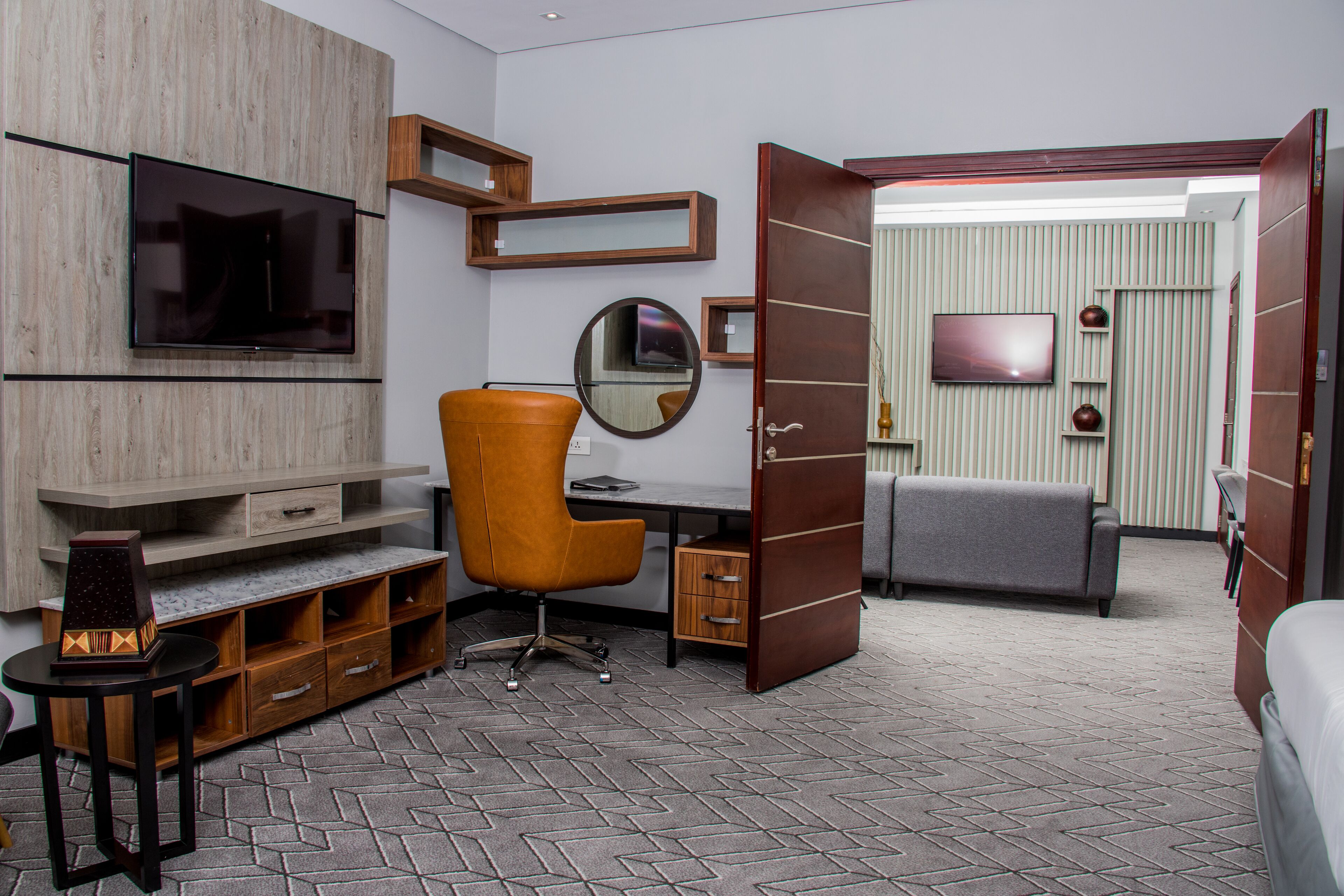 A modern hotel room with a wooden desk, an orange chair, a flat-screen TV, a round mirror, and a grey sofa.
