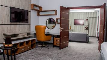 A modern hotel room with a wooden desk, an orange chair, a flat-screen TV, a round mirror, and a grey sofa.