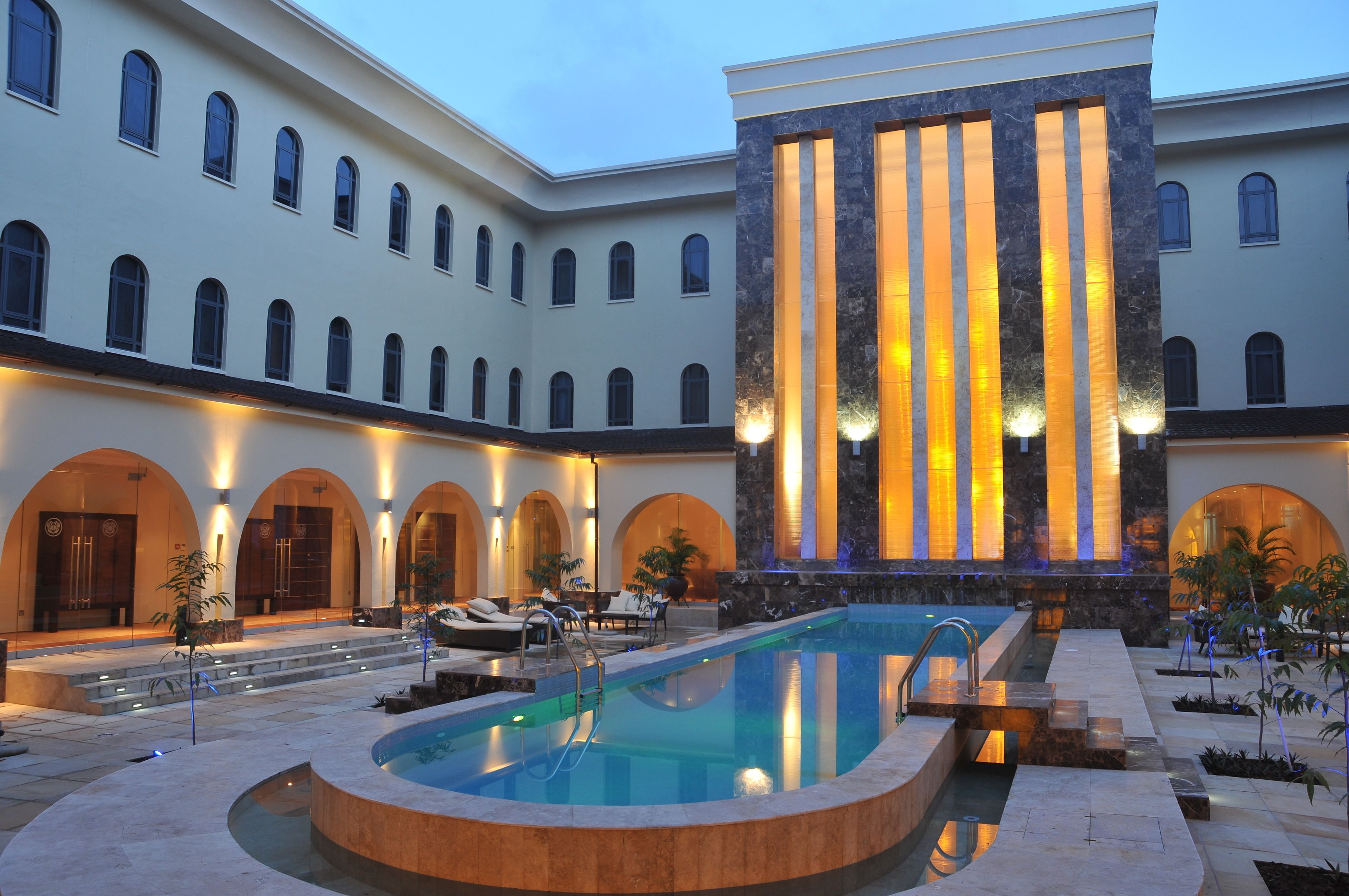 A luxurious hotel courtyard with a central swimming pool, multiple arches, and a large building with vertical lighting.