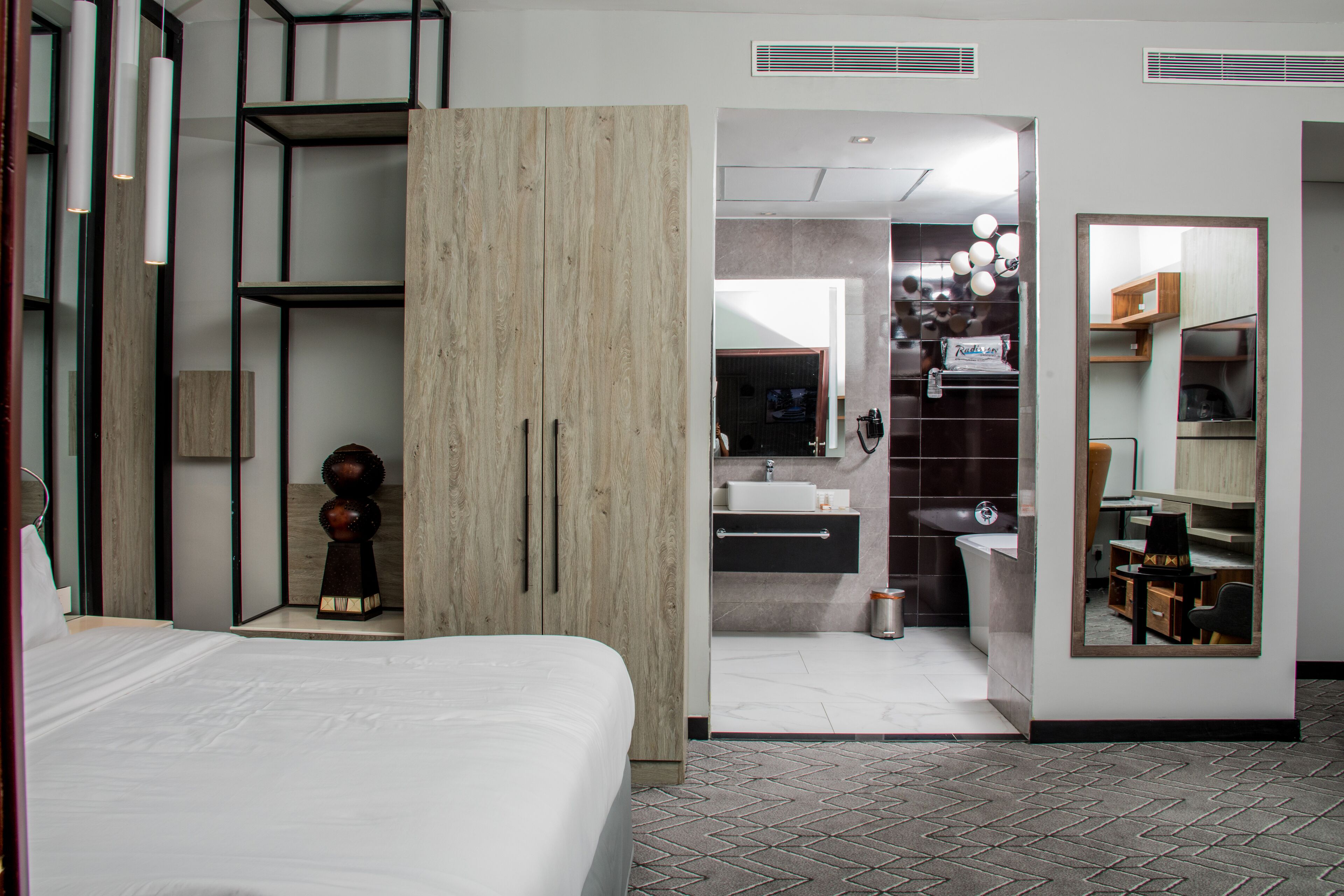 A modern hotel room with a bed, a wooden wardrobe, a bathroom with a sink and mirror, and a small seating area.