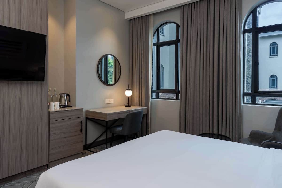 A hotel room with a bed, a desk with a chair, a TV, a mirror, and a window with curtains.
