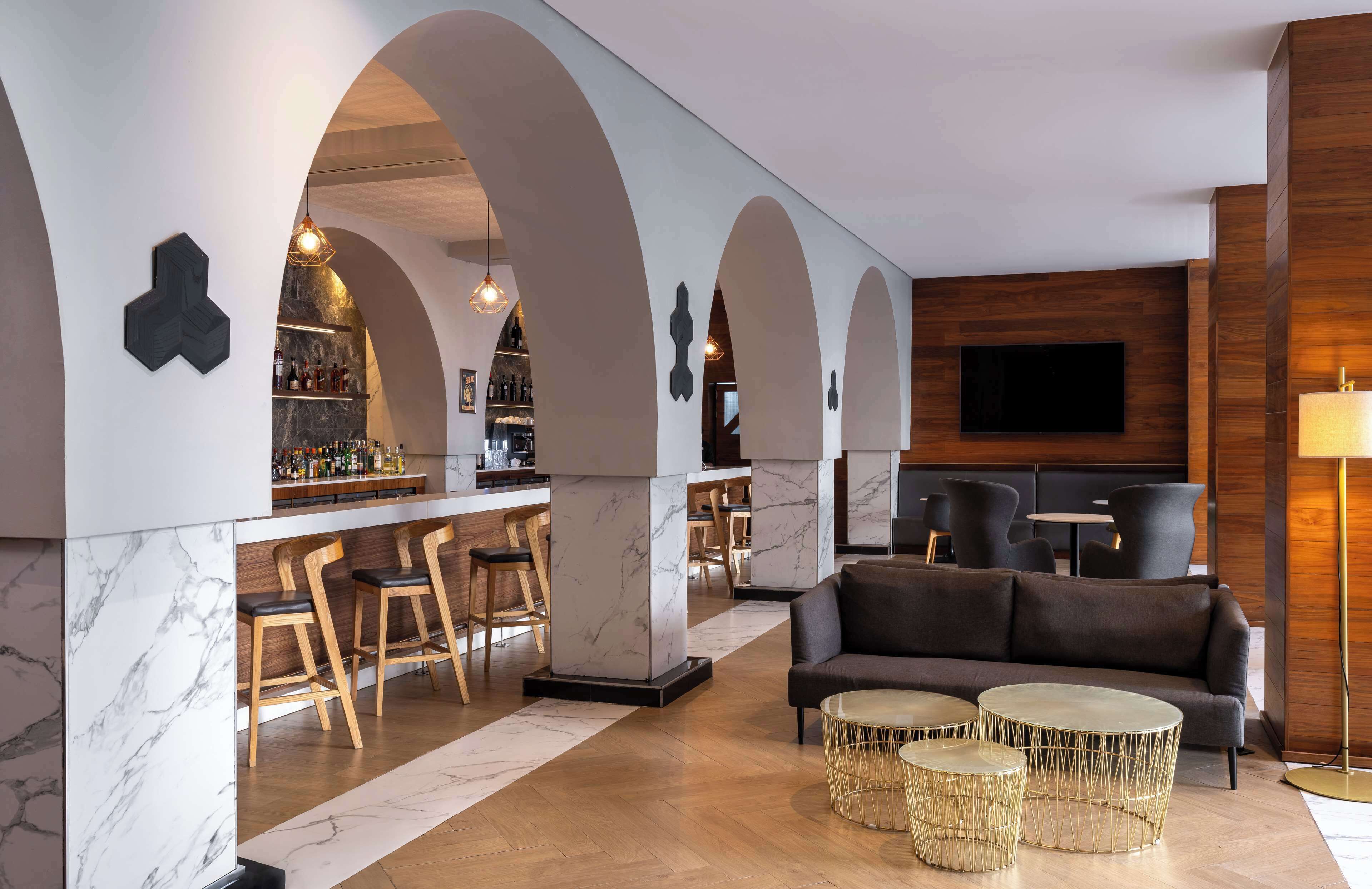 A modern bar area with marble countertops, wooden bar stools, and a seating area with a sofa and round tables.