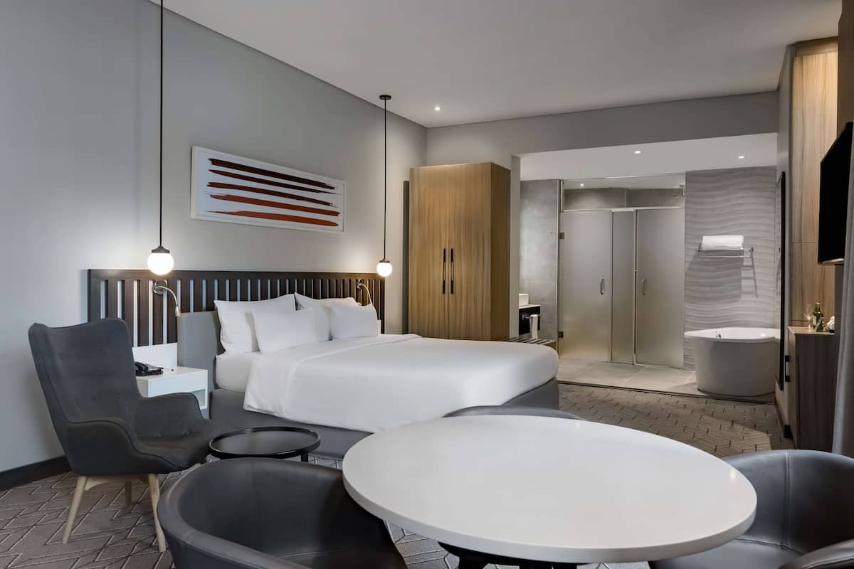 A modern hotel room with a bed, a seating area with a round table, a wardrobe, and a bathroom with a bathtub.