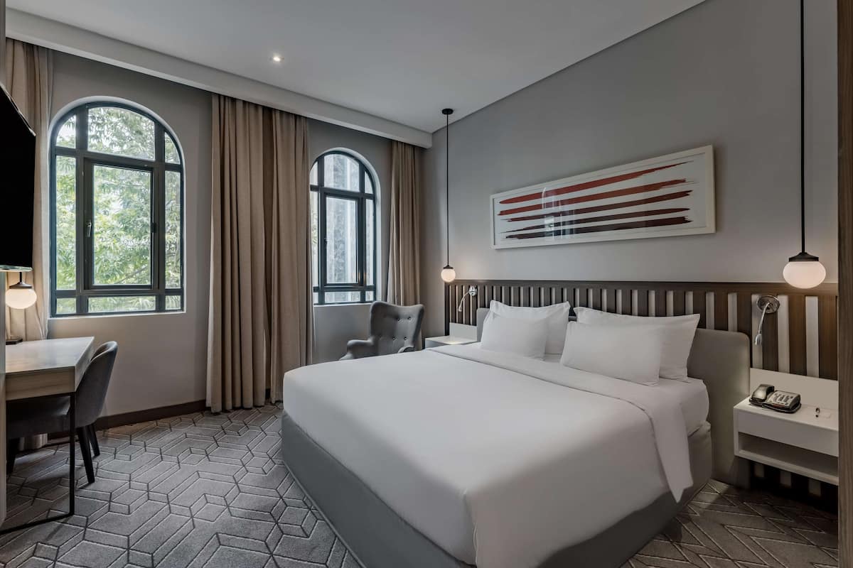 A modern hotel room with a large bed, a desk with a chair, a small table, and a nightstand with a lamp.