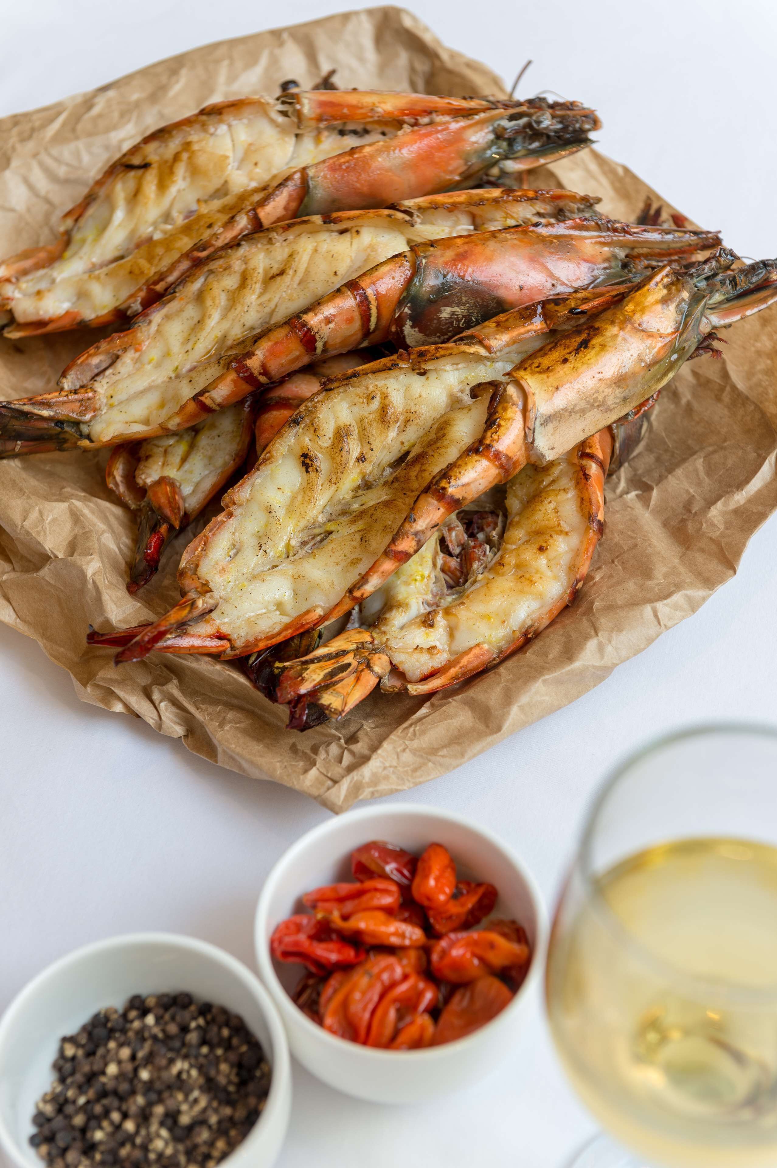 Gambas a la parrilla en una bandeja forrada con papel, junto con recipientes pequeños de pimienta negra y chiles rojos, y una copa de vino blanco.
