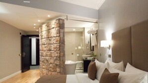 Et moderne hotelværelse med en stenvæg, en stor seng med hvide og brune puder, et sengebord med en lampe, og et badeværelse, der kan ses gennem en glasdør.