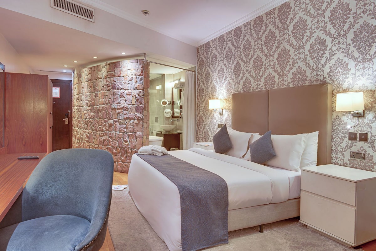 A hotel room with a stone accent wall, a bed with a headboard, bedside tables, a desk, and a chair.