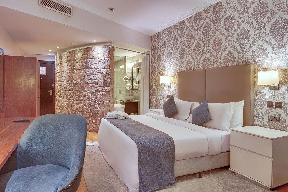 A hotel room with a stone accent wall, a bed with a headboard, bedside tables, a desk, and a chair.