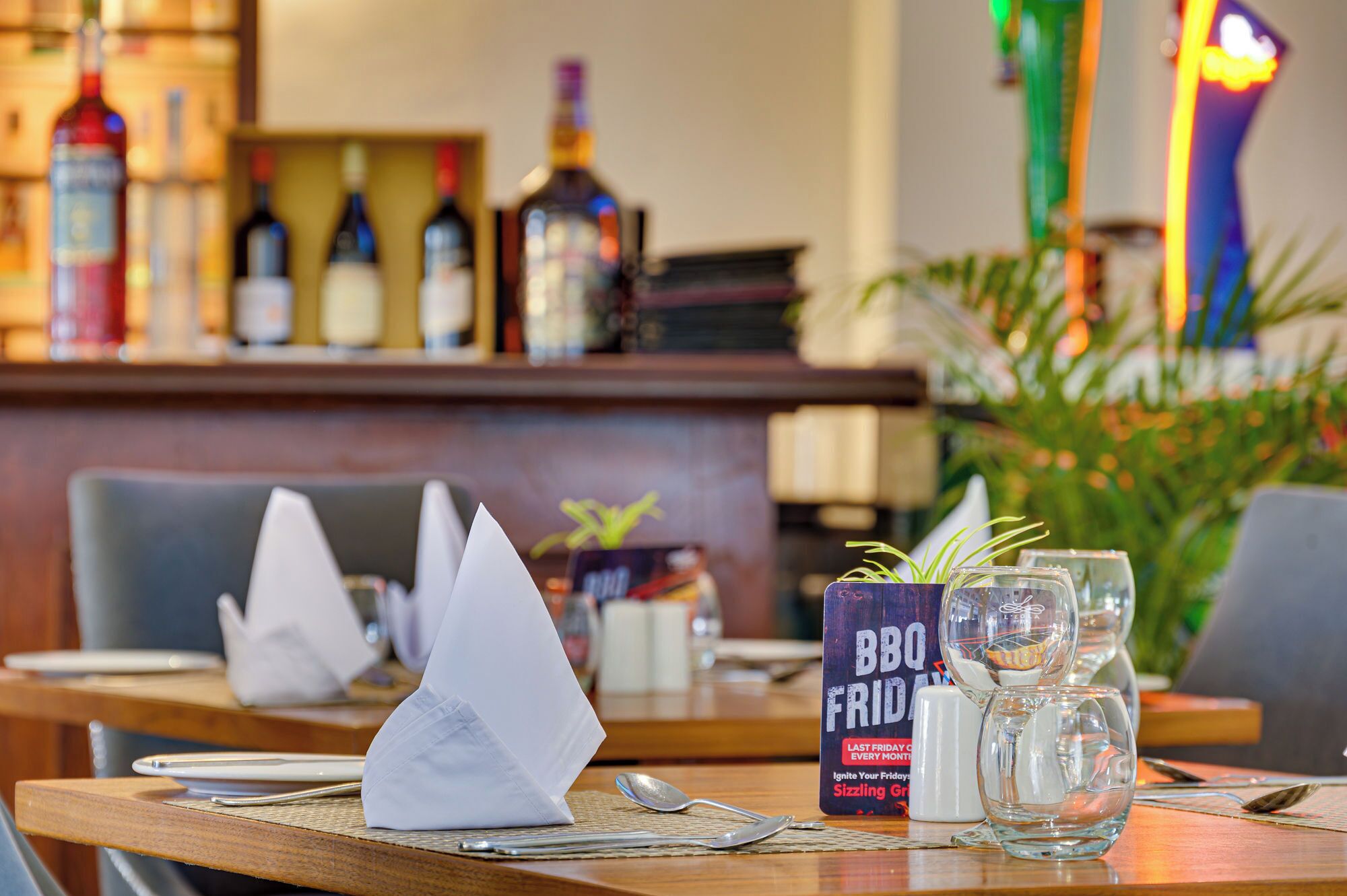 Un ambiente de restaurante con una zona de bar al fondo, un atril para el menú y una mesa con servilleta, cuchara y vasos.