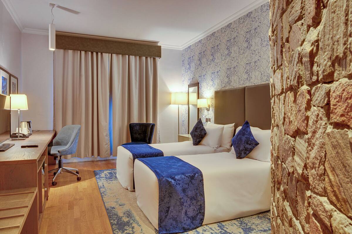 A hotel room with a bed, two bedside tables, a desk with a chair, a lamp, and a large window with curtains.