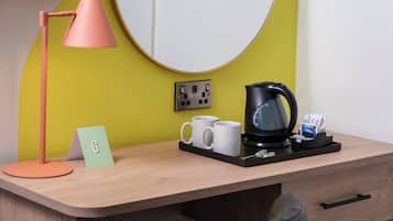 Ein Hotelzimmer mit Schreibtisch aus Holz, einer schwarzen Kaffeemaschine, zwei weißen Tassen und einer rosa Schreibtischlampe. Auf dem Schreibtisch liegt eine kleine Karte.