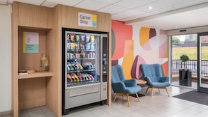 A vending machine labeled 'Garner Shop' with snacks and drinks, a small shelf with glassware, and two blue chairs in a modern interior.