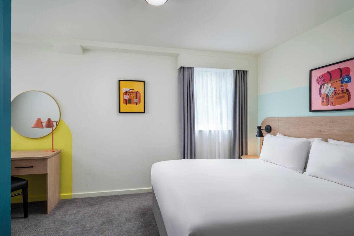 A hotel room with a bed, a desk, a chair, a round mirror, a framed picture, and a window with curtains.
