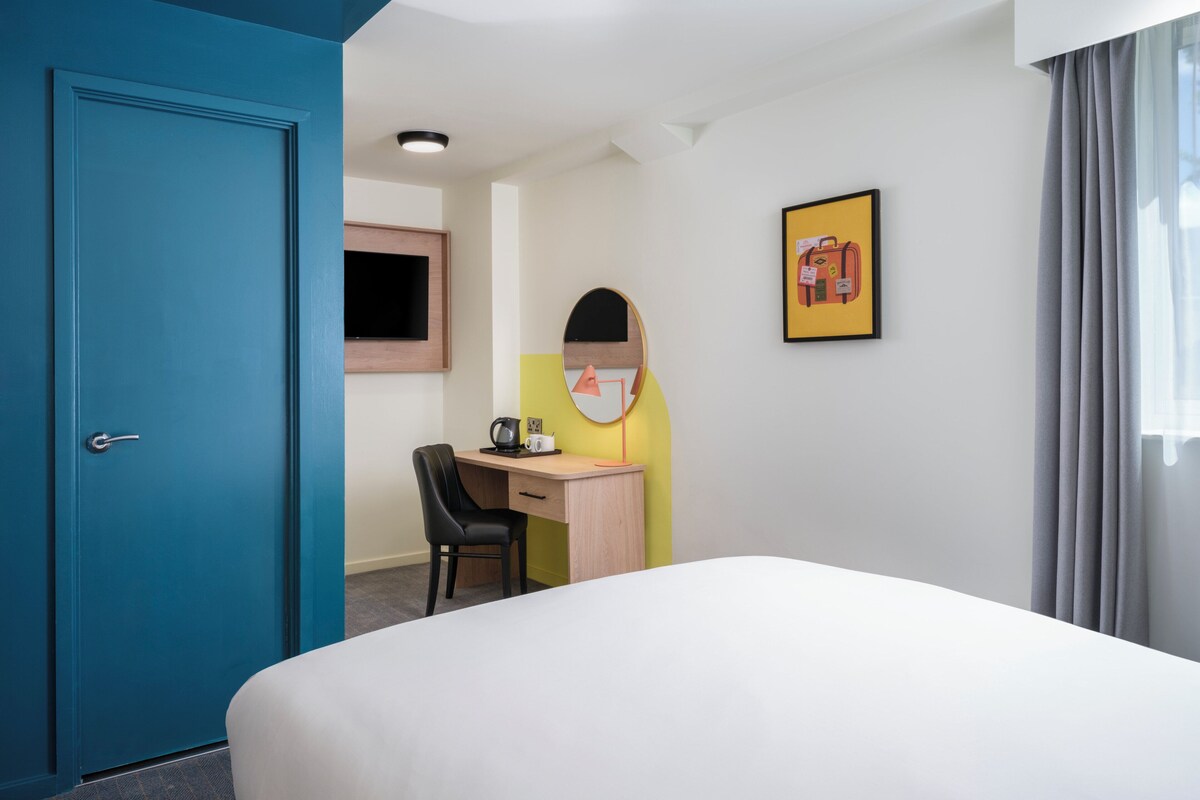 Une chambre d’hôtel avec un lit, un bureau, une chaise, une télévision et un tableau encadré au mur.