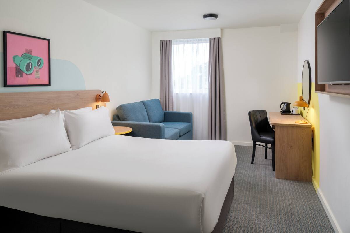A hotel room with a large bed, a desk with a chair, a small table, a TV, and a window with curtains.
