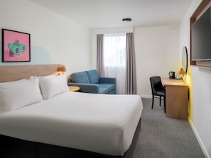 Ett hotellrum med en stor säng, ett skrivbord med en stol, ett litet bord, en tv och ett fönster med gardiner.