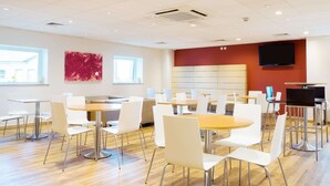 A modern dining area with round tables and white chairs, a red accent wall, and a flat-screen TV mounted above a seating area. - Travelodge Middlesbrough (Middlesbrough)