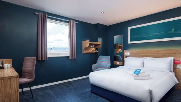A hotel room with a bed, a desk, a chair, a window with curtains, a painting on the wall, and a view of the sky. - Travelodge London Barking (Barking)
