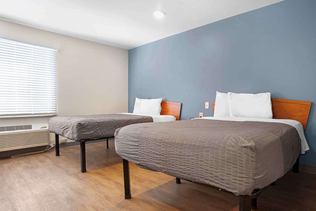 A hotel room with two single beds, a large window with blinds, a wall-mounted heater, and a blue accent wall.