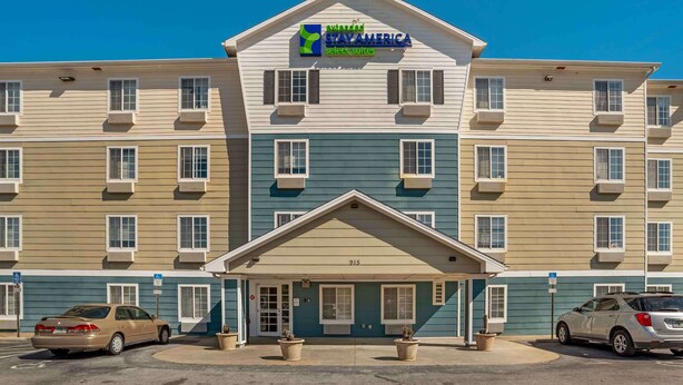 A multi-story hotel with a central entrance, air conditioning units, and a parking area with two cars. - Extended Stay America Select Suites - Fort Walton Beach (Fort Walton Beach)
