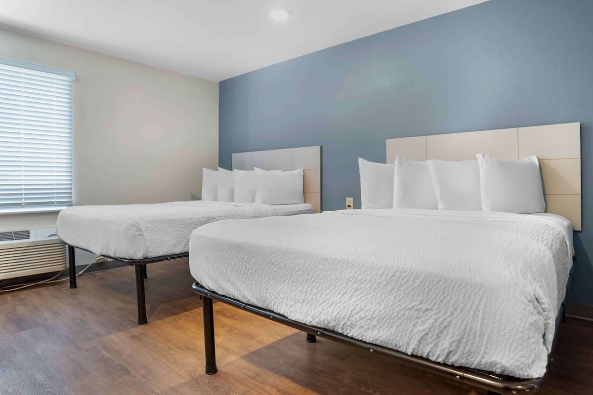 A hotel room with two beds, a large window with blinds, and a wall with a blue and white pattern.