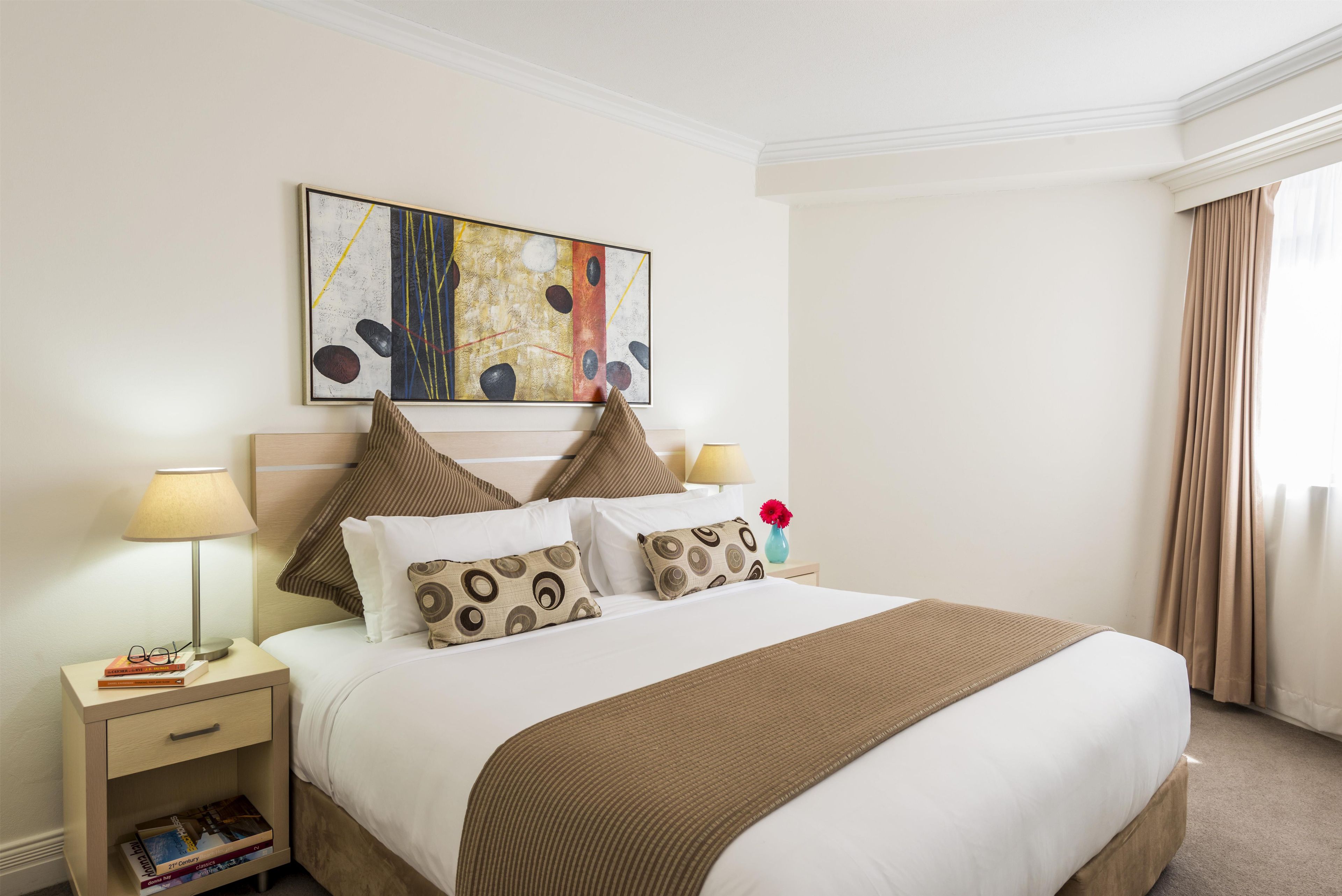 Una habitación de hotel con cama, almohadas, mesita de noche, lámpara, un cuadro y una ventana con cortinas.