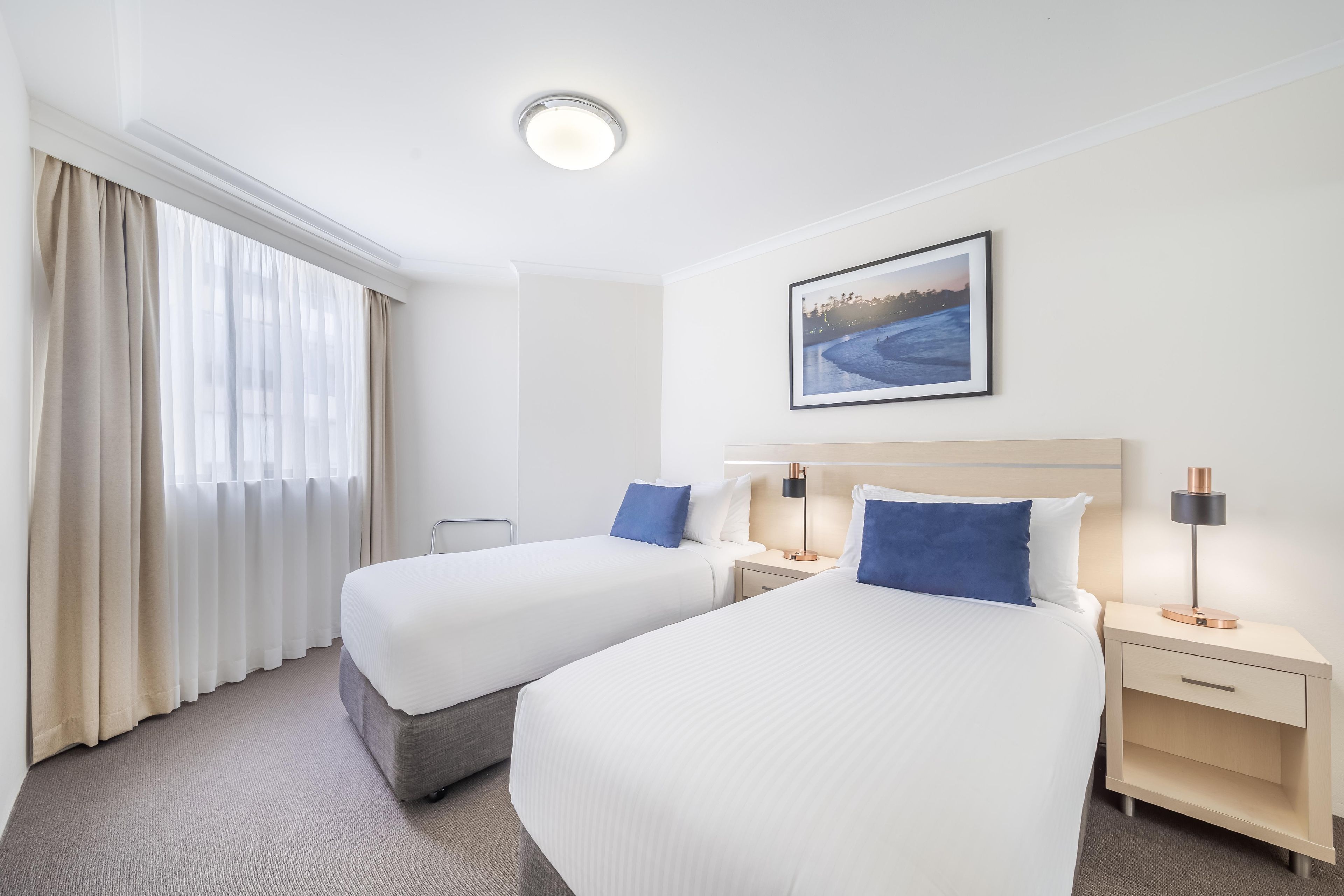 A hotel room with two beds, a nightstand with a lamp, a framed picture on the wall, and a window with curtains.